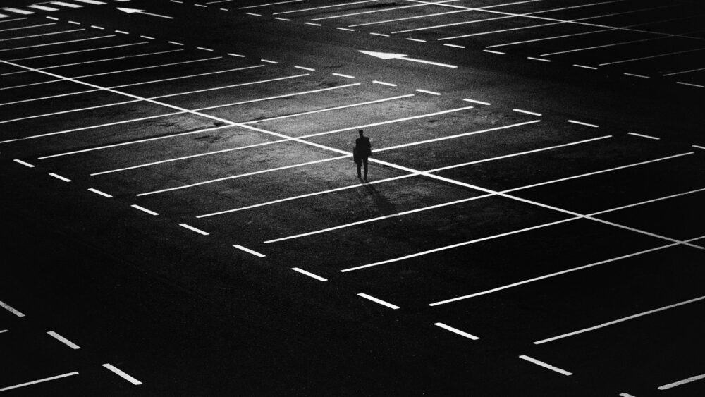 Black and white photo of empty parking lot with no cars and one person walking through it