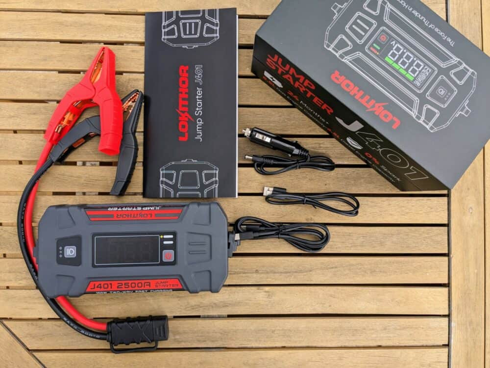 Lokithor J401 portable jump starter on a wooden table, with the product box alongside. The box contents are  arranged around it: three charging cables, an instruction booklet, and a set of short jumper leads.
