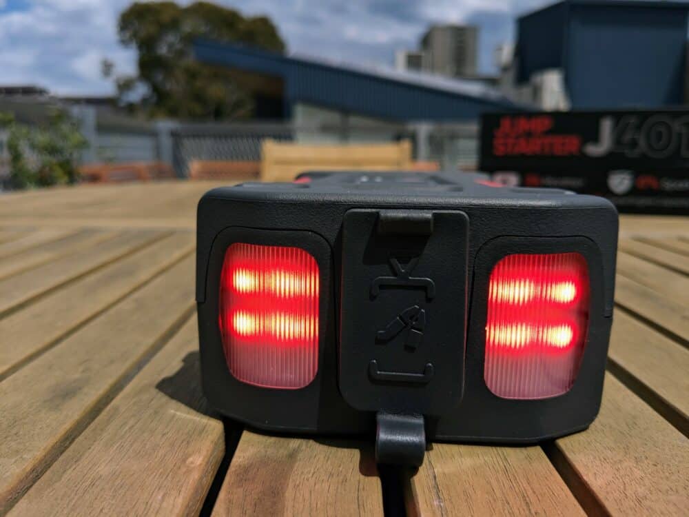 Front-on view of the Lokithor J401 portable jump starter sitting on a wooden table, with red LEDs shining through two transparent panels. Product box (blurred) visible in the background.