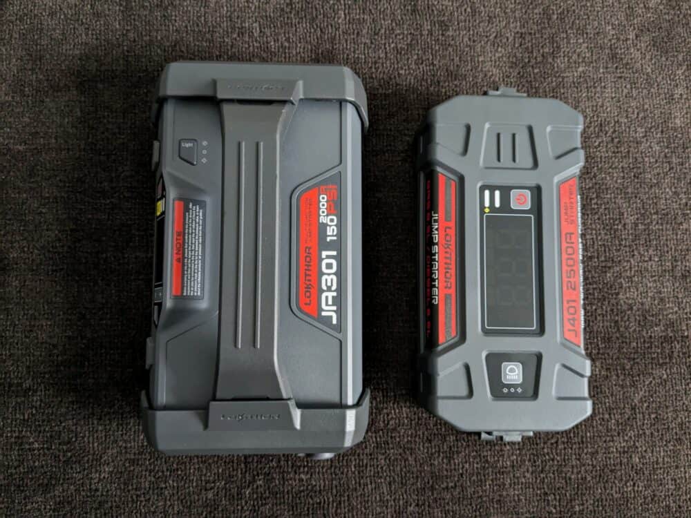 Two portable jump starters sitting on dark carpet. The larger one on the left is the model number JA301, and a wide handle. The smaller one on the right has the model number J401, and a screen on top but no handle.