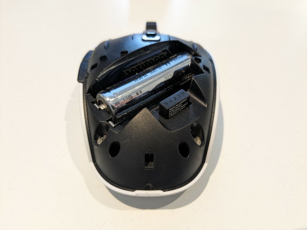 RAZER Orochi mouse with top cover removed, on white table. Visible inside the mouse is a single AA battery, and the top part of a USB dongle in a small compartment.