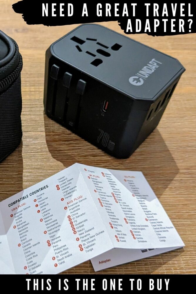 Black travel adapter and partial view of the instruction booklet and travel case for it, all sitting on a wooden table. Text overlaid: "Need a great travel adapter?" at top and "This is the one to buy" at the bottom.