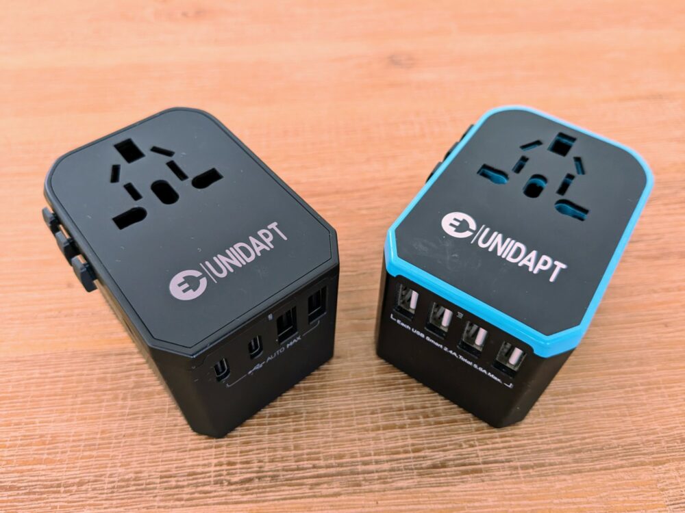 Two Unidapt international travel adapters beside each other on a wooden table. The new model on the left is entirely black and has two USB C and two USB-A ports visible on the base, the old model on the right has a blue border and four USB-A ports on the base.