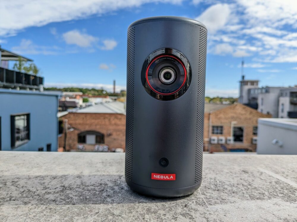 Nebula Capsule 3 Laser cylindrical portable speaker sitting on a concrete wall with blurred buildings and sky visible behind
