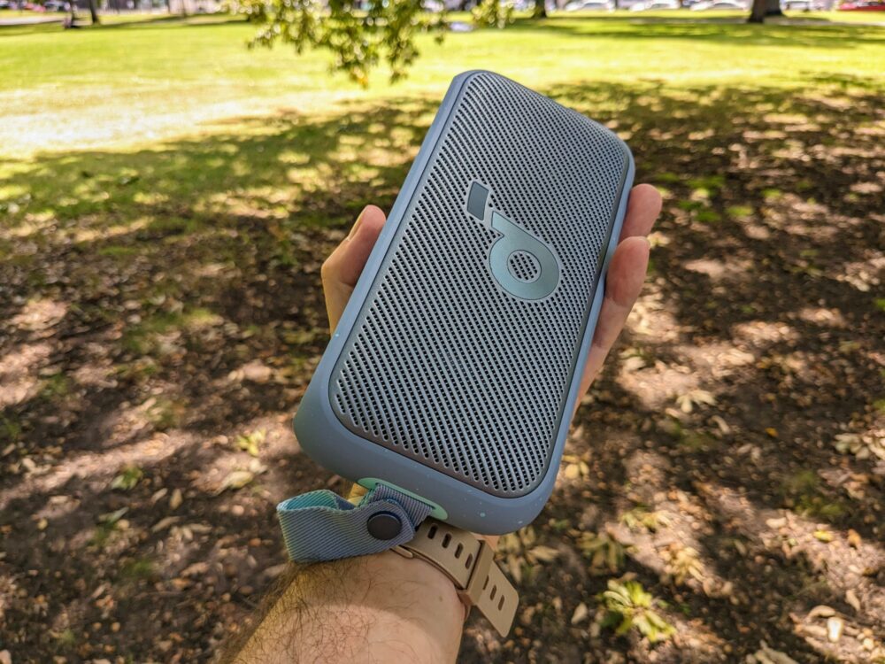 An outstretched arm and hand holding a portable speaker, with dirt, grass, and trees blurred in the background.