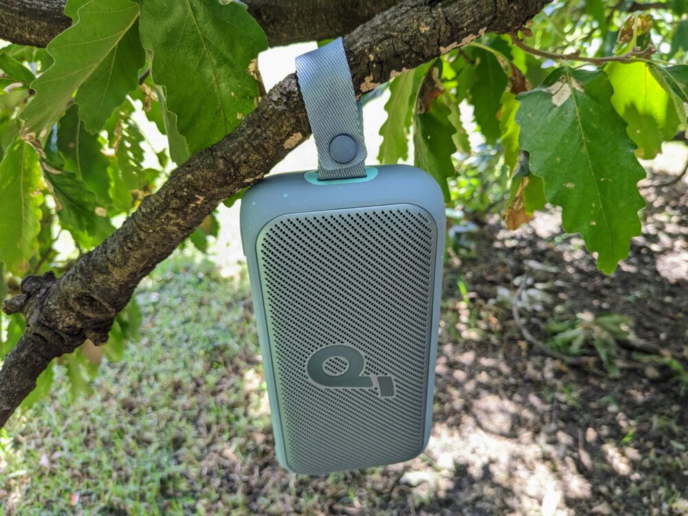 Portable speaker hanging by its strap from the branch of a tree with dirt and leaves behind.