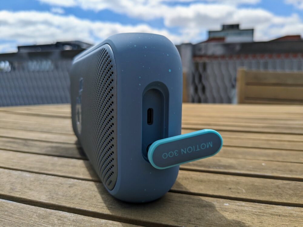 Portable speaker sitting on a wooden outdoor table with blurred buildings in the background. The flap covering the charging socket is open, displaying the USB C port behind it.