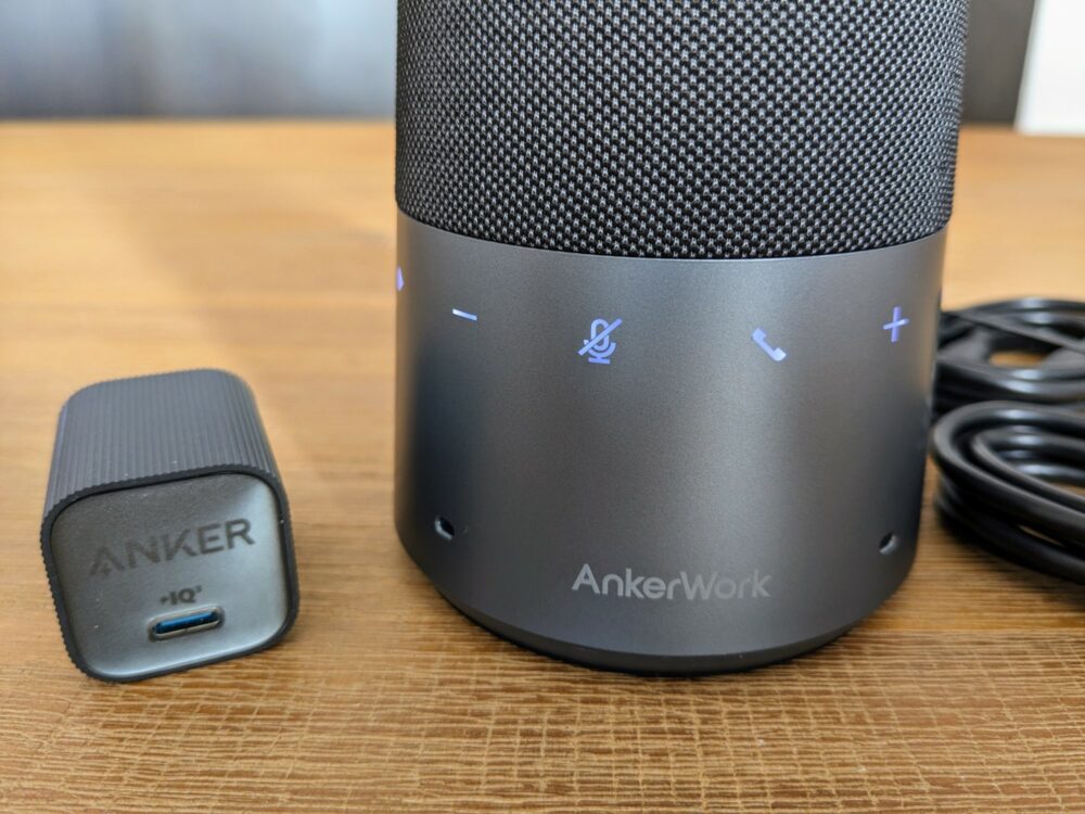 Closeup view of the bottom section of a cylindrical speakerphone with several illuminated buttons and an AnkerWork logo. A small USB C wall charger sits alongside, plus two black cables