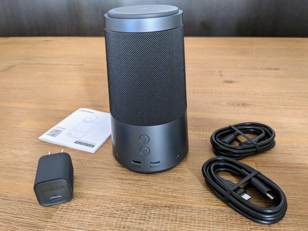 Cylindrical speakerphone standing upright on table, with a wall charger, two USB C cables, and an instruction manual alongside