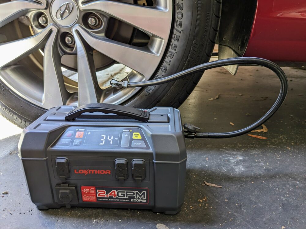 Portable jump starter unit on the floor of a garage, connected by a hose to a car tire. Status screen reads 34 PSI.