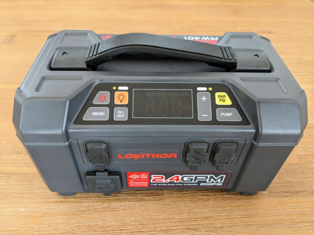 Lokithor portable jump starter sitting on a wooden table, with several ports covered by rubber stoppers and a status screen (turned off) on top.