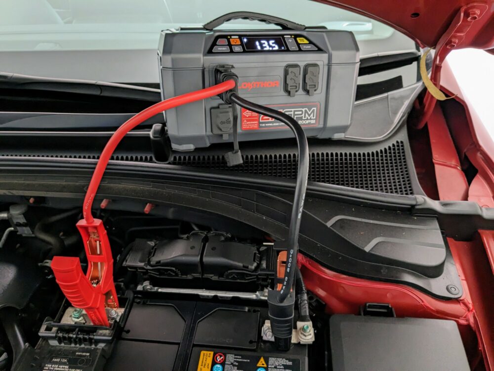Portable jump starter with black and red jumper cables connected to both it and a car battery. Screen on jump starter shows 13.5 volts.