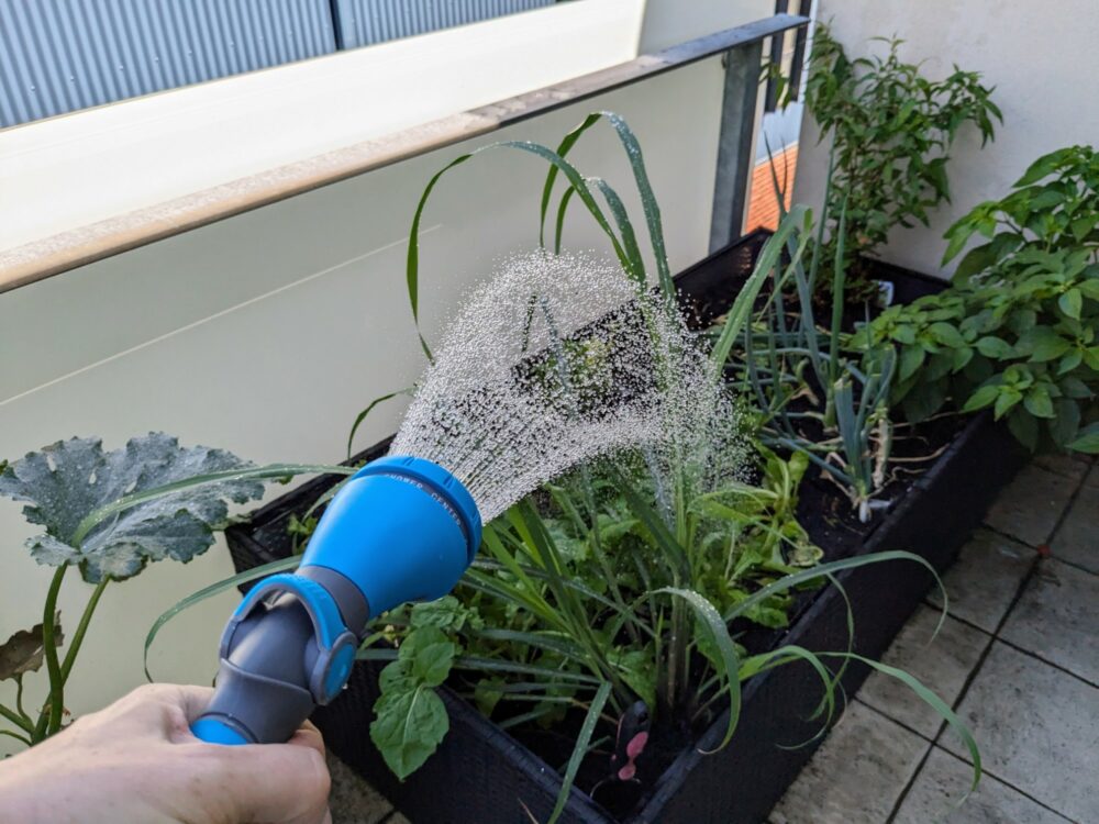 Hand holding a hose attachment that is spraying a shower of water onto a planter box of vegetables on a balcony.