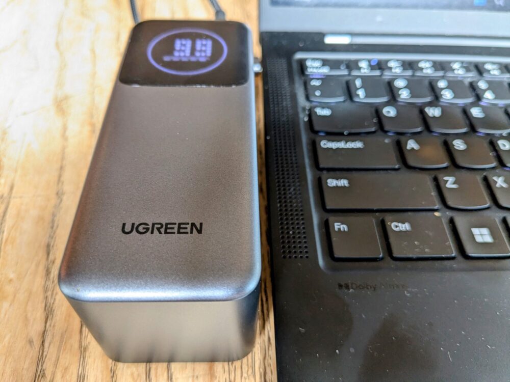 Power bank sitting on a wooden table, plugged into a black laptop that is sitting alongside. 99% remaining capacity is showing on the power bank display