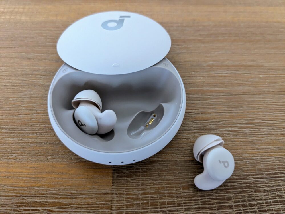 Circular earbud case open on a wooden table, with one earbud inside and the other sitting alongside