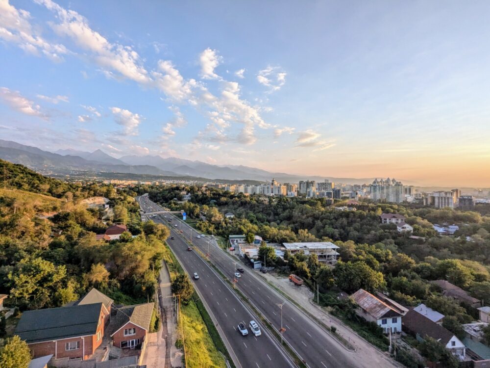 Aerial view of a multi-lane road with many trees on either side, a built-up section of a city to the right, and mountains in the distance, taken at sunset