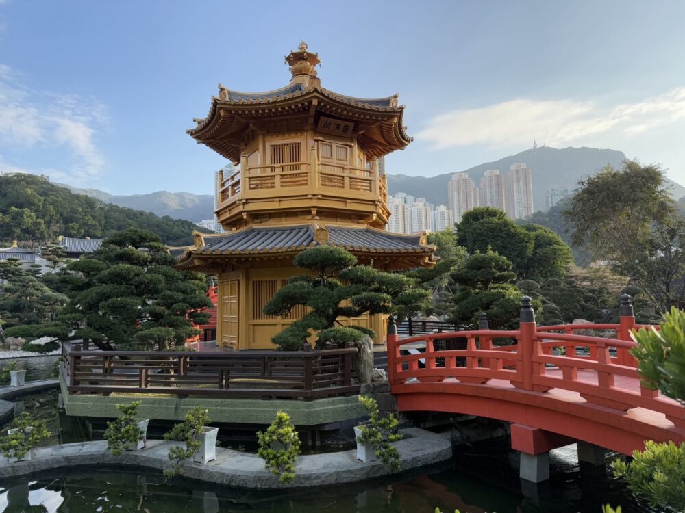 Chinese-style temple in a park with red wooden bridge leading to it and skyscrapers visible in the background