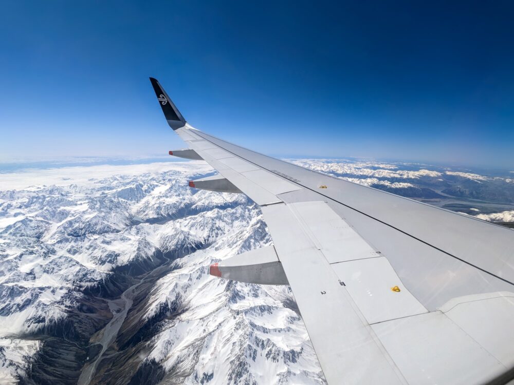 Wing of an Air New Zealand plane over a snow-capped mountain range with a river valley below