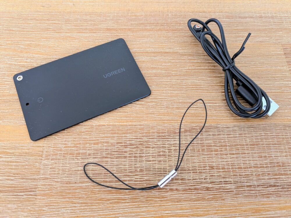 Slim black tracking device with UGreen branding on a wooden table, beside a charging cable and lanyard